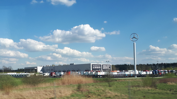 Eine Mercedes-Benz-Fabrik in Deutschland mit Fahrzeugen auf der Straße, umgeben von Gras, Pflanzen, Bäumen, Gebäuden und Polen, von denen einer ein Logo trägt, unter einem bewölkten Himmel.