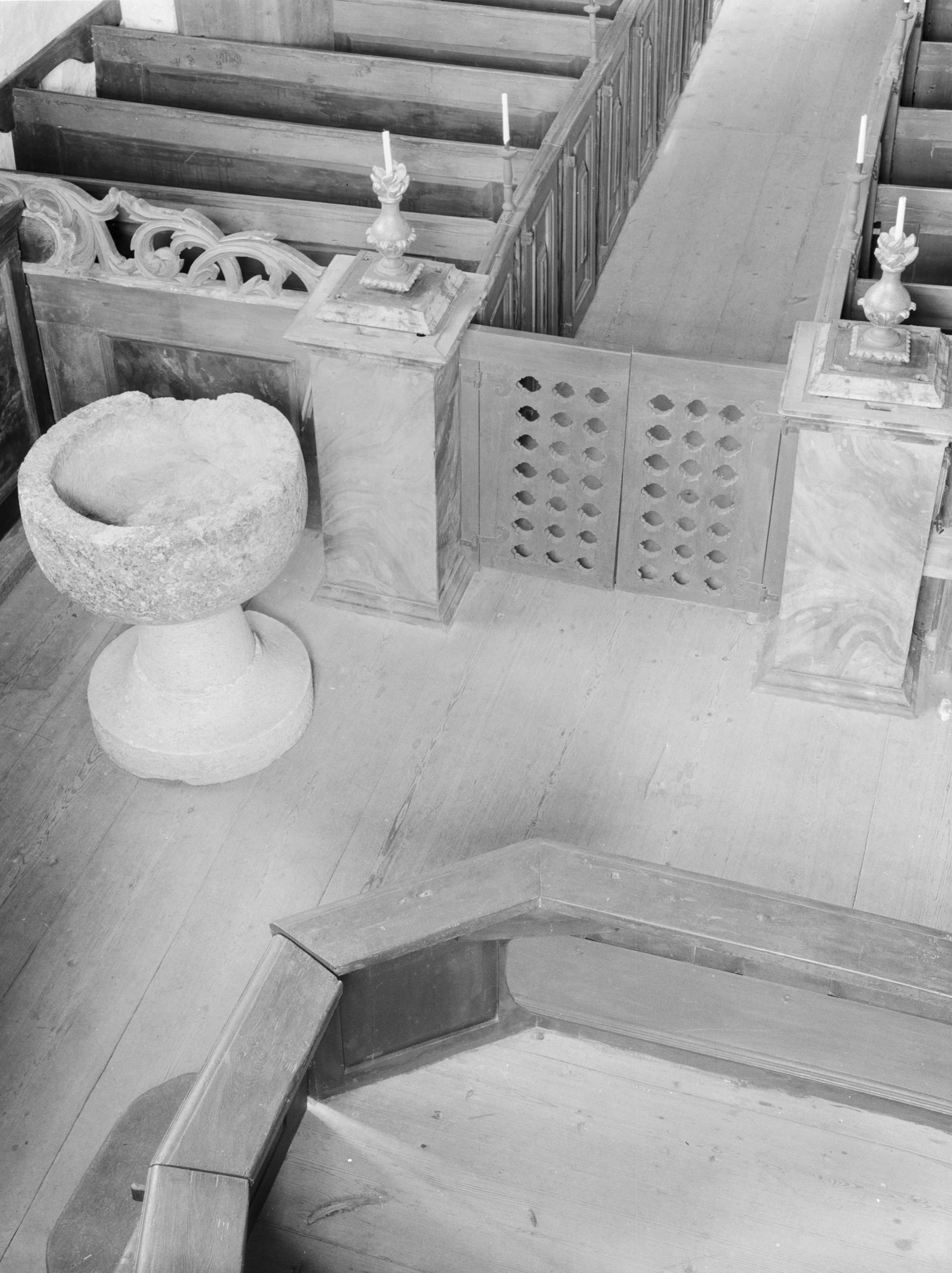 Ein Schwarz-Weiß-Foto einer Kircheninnenansicht mit hölzernen Kirchenbänken, die in Reihen angeordnet sind, einem steinernen Becken in der Mitte und Kerzen auf Ständern auf beiden Seiten des Beckens.