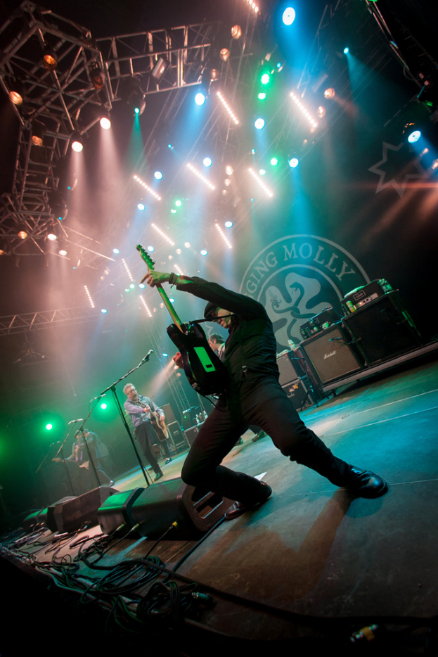 Ein musikalisches Konzert mit drei Personen auf der Bühne: zwei spielen Gitarren rechts mit Mikrofonen davor, ein Hintergrund mit "MOLLEY" geschrieben und zwei Schlangenlogos, sowie Bühnenlichter oben.