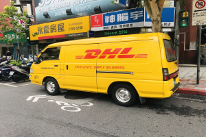 Ein gelber DHL-Lieferwagen auf einer Straße neben Gebäuden, Bäumen, Schildern, Polen, Verkehrszeichen und einem Fußgänger.
