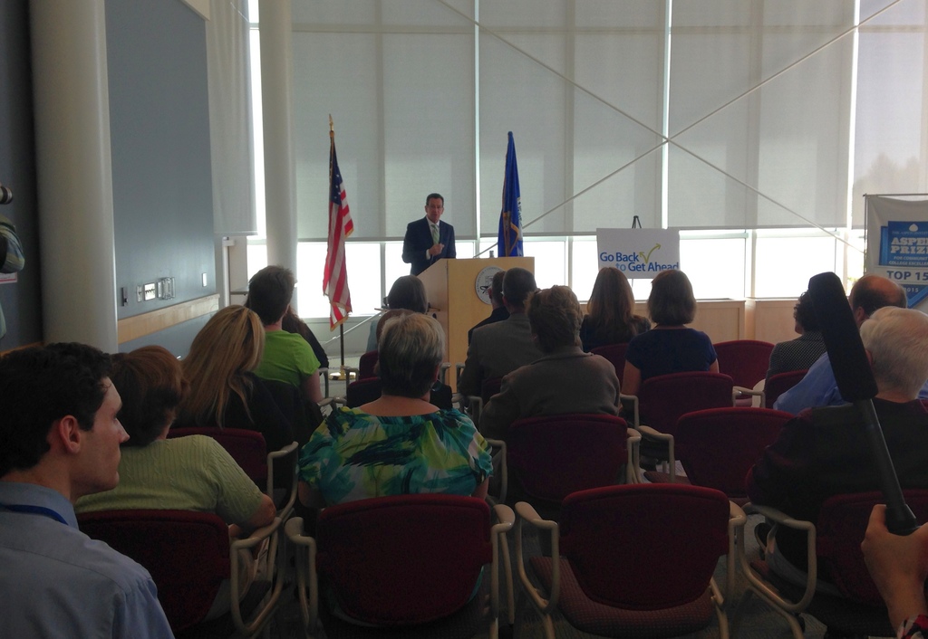 Ein Mann steht an einem Rednerpult und spricht, mit zwei Fahnen daneben, während viele Menschen auf Stühlen sitzen, mit Säulen und Fenstern im Hintergrund.