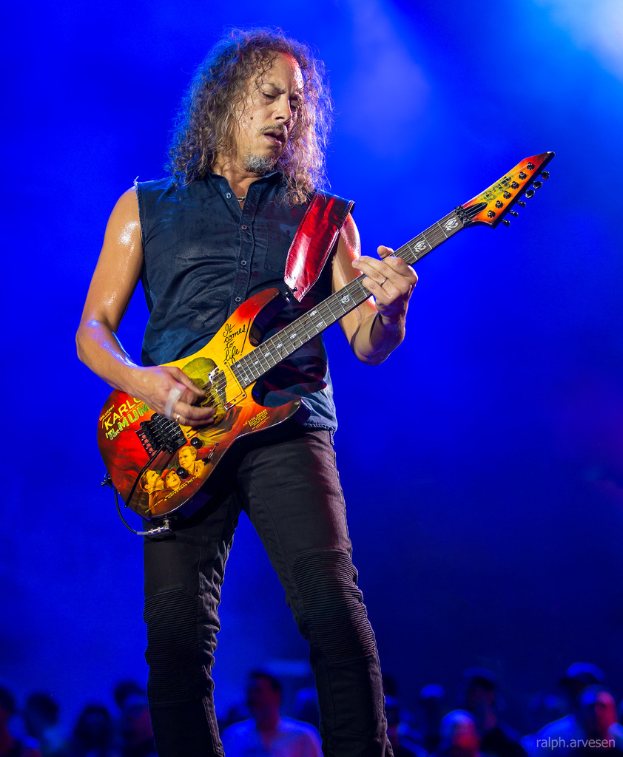 Eine Person in einem schwarzen Kleid steht in der Mitte eines musikalischen Konzerts und spielt eine Gitarre, während viele Menschen von unten zuschauen.