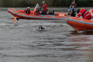 Eine Gruppe von Menschen in einem aufblasbaren Boot auf einem Fluss, mit zwei Personen im Wasser im Vordergrund und einigen Pflanzen im Hintergrund, alle mit Schwimmwesten und Helmen ausgestattet, was auf einen Rettungseinsatz hinweist.
