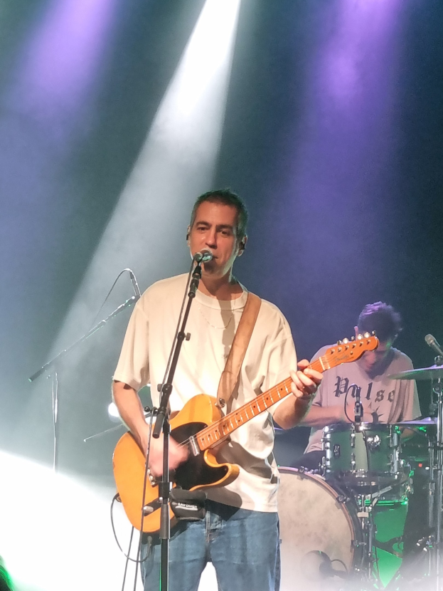 Ein Mann spielt eine E-Gitarre auf der Bühne, während er in ein Mikrofon singt, mit einem anderen Musiker, der im Hintergrund Schlagzeug spielt.