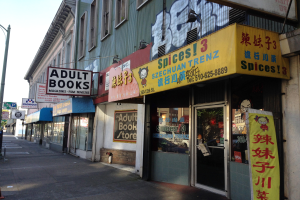 Eine belebte Straße mit Gebäuden, Fahrzeugen, Bäumen und einem Schaufenster mit der Aufschrift "Erwachsene Bücher" unter einem klaren Himmel.