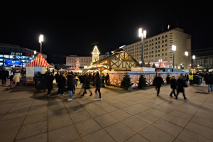 Ein belebter Stadtplatz bei Nacht, erhellt von Stra├čenlaternen und Gebäudelichtern, mit Menschen, Ständen und Bäumen, unter einem dunklen Himmel.