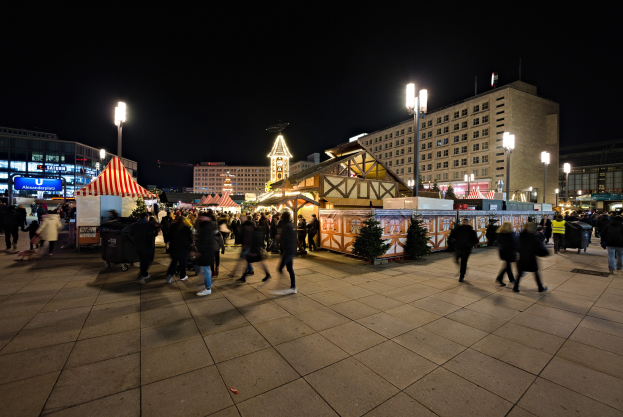 Ein belebter Stadtplatz bei Nacht, erhellt von Stra├čenlaternen und Gebäudelichtern, mit Menschen, Ständen und Bäumen, unter einem dunklen Himmel.