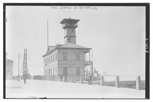 Schwarz-weiß-Foto einer Feuerwache in Battery, New York, mit Menschen, Pfählen, einem Zaun, Booten auf dem Wasser und einem bewölkten Himmel.