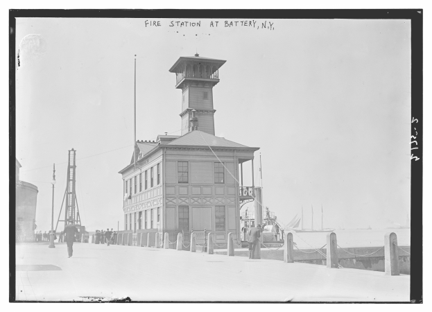 Schwarz-weiß-Foto einer Feuerwache in Battery, New York, mit Menschen, Pfählen, einem Zaun, Booten auf dem Wasser und einem bewölkten Himmel.